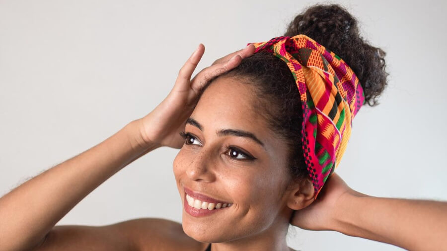 Eine junge schöne Frau trägt ein traditionelles farbenfrohes afrikanisches Kente-Headwrap von Curly N Covered. Sie lächelt und sieht glücklich aus.