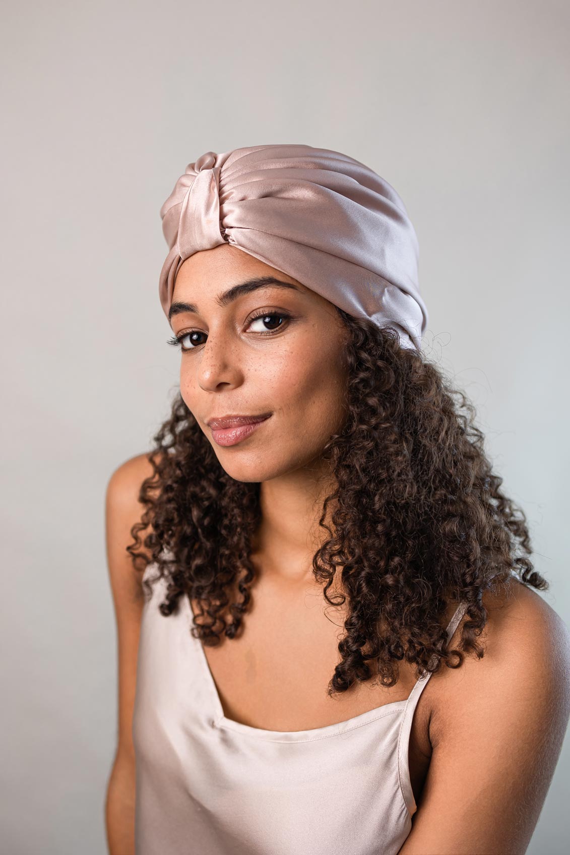 Wunderschöne Frau mit dunklen lockigen Haaren trägt einen sand-farbenen Seidenturban aus reiner Maulbeerseide von CURLY N COVERED zum Schutz ihrer Locken